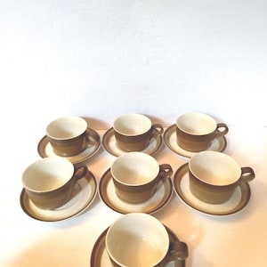 May include: A set of seven ceramic tea cups and matching saucers. Each cup features a speckled cream interior and a two-tone exterior with a dark brown base and a lighter tan band. The saucers have a matching brown and tan circular design.