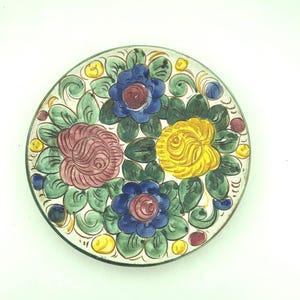May include: A round, hand-painted ceramic plate featuring a floral design. The flowers are in shades of blue, yellow, and pink, surrounded by green leaves and yellow accents. The plate has a white background and a dark rim.
