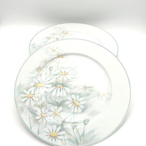 May include: Two round white ceramic dinner plates featuring a delicate watercolor-style illustration of white daisy flowers with yellow centers and soft green foliage. Each plate has a thin, light green rim around the outer edge.