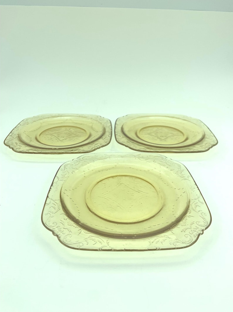 Puede incluir: Tres platos cuadrados de color &aacute;mbar con un patr&oacute;n floral decorativo en los bordes. Cada plato tiene un c&iacute;rculo central ligeramente elevado. Los platos son de vidrio y est&aacute;n dispuestos sobre una superficie blanca.