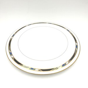 Puede incluir: Un plato redondo de porcelana blanca con un patrón decorativo en el borde. El borde está adornado con formas geométricas en negro, dorado y azul, acentuadas por finas líneas grises. El centro es blanco liso con una fina línea circular dorada.