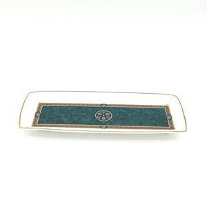 May include: A rectangular white ceramic tray featuring a central dark teal marbled panel. The panel is bordered by a geometric gold and black pattern, with a circular emblem in the center containing four small teal shapes and gold accents.