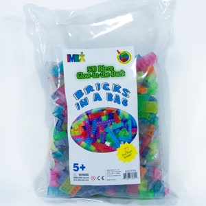 May include: A clear plastic bag filled with 500 glow-in-the-dark building bricks. The bag has a label that says "500 Piece Glow-in-the-Dark Bricks in a Bag". The label also says "27 Roof Pieces Included!" and "5+".