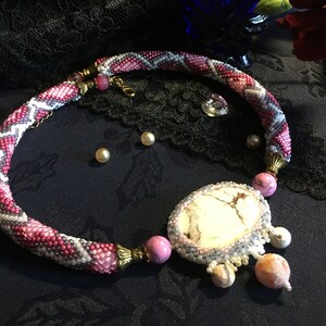 Könnte beinhalten: Eine rosafarbene, weiße und graue Perlenkette mit einem großen weißen Steinanhänger und kleineren rosa und orangefarbenen Steinen. Die Kette liegt auf einem schwarzen Stoffhintergrund.