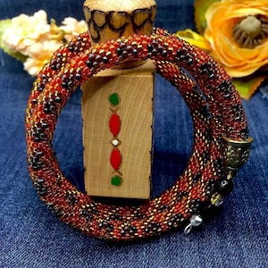 May include: A close-up of a beaded bracelet made with small, colourful beads in shades of orange, brown, and gold. The bracelet is wrapped around a wooden block and has a decorative metal clasp.