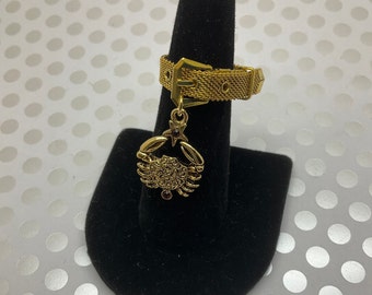 May include: Gold-tone ring with a mesh band and a dangling charm of a crab with a red gemstone. The ring is displayed on a black velvet ring holder.