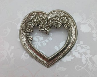 May include: A silver heart-shaped brooch with a floral design. The flowers are arranged around the open heart shape.