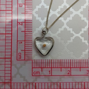 May include: A silver heart-shaped pendant necklace with a clear glass insert containing a small, light brown seed. The necklace is measured against a ruler in centimetres.