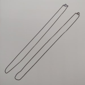 May include: Two silver chains with lobster claw clasps. The chains are laid out on a white surface.