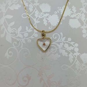 May include: A gold chain necklace with a heart-shaped pendant. The pendant is clear glass and contains a small, brown seed inside.