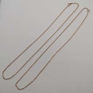 May include: Two delicate gold-coloured chain necklaces with small links are displayed against a white background. The chains are laid out diagonally, showcasing their length and design. The necklaces are likely for jewellery.