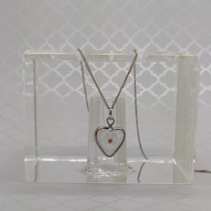 May include: A silver heart-shaped pendant necklace with a small, round, light brown seed inside. The pendant is hanging from a silver chain and is displayed in a clear acrylic box.