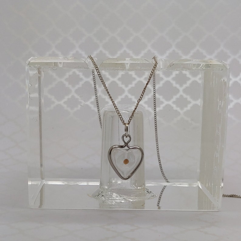 May include: A silver heart-shaped pendant necklace with a small, round, light brown seed inside. The pendant is hanging from a silver chain and is displayed in a clear acrylic box.