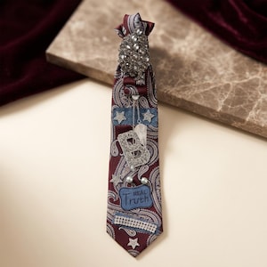 May include: A burgundy paisley tie with star and letter embellishments. The tie features a jeweled top, denim patches, and the words "Real Truth" on a blue patch. The tie is decorated with rhinestones and pearls.