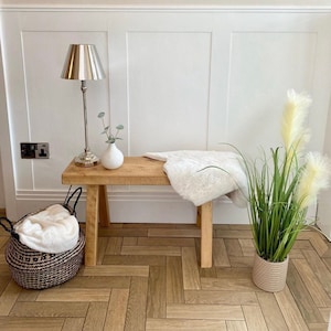 Puede incluir: Un banco de madera con una manta de piel de oveja blanca, una l&aacute;mpara plateada, un jarr&oacute;n blanco con vegetaci&oacute;n, una cesta tejida con una manta blanca y una planta en maceta con penachos altos y plumosos. La habitaci&oacute;n tiene una paleta de colores neutros.