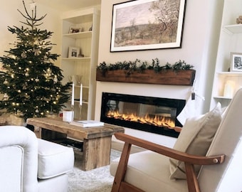 May include: A white living room with a Christmas tree, a fireplace, a wooden coffee table, and a white armchair with a beige pillow. The fireplace has a black electric fire insert and a wooden mantel with greenery.