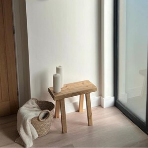 Puede incluir: Un peque&ntilde;o taburete de madera natural con tres patas. Dos velas blancas en forma de pilar est&aacute;n encima. Una cesta tejida con una manta crema est&aacute; en el suelo. La habitaci&oacute;n es luminosa, con una puerta de cristal.