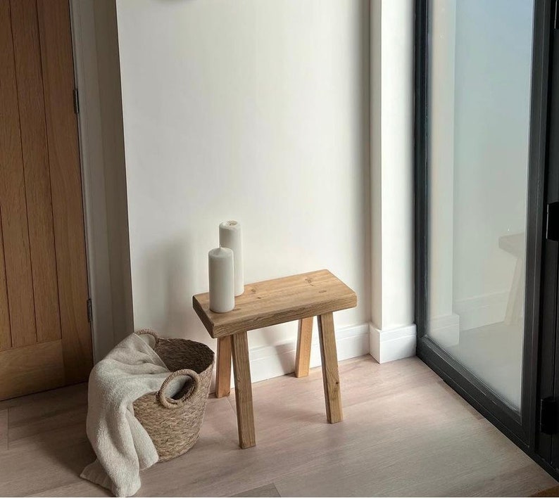 Puede incluir: Un peque&ntilde;o taburete de madera natural con tres patas. Dos velas blancas en forma de pilar est&aacute;n encima. Una cesta tejida con una manta crema est&aacute; en el suelo. La habitaci&oacute;n es luminosa, con una puerta de cristal.