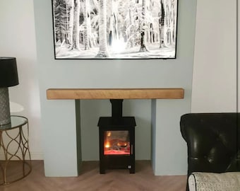 May include: A black and white framed print of a snowy forest scene hangs above a wooden mantel. Below, a black fireplace with a glowing fire effect is set into a light blue wall. A black lamp and a side table are visible.