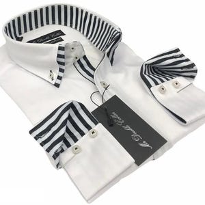 Puede incluir: Camisa blanca con cuello y puños a rayas blancas y negras. La camisa tiene cuello abotonado y puños abotonados. Una etiqueta negra con texto blanco está adjunta.