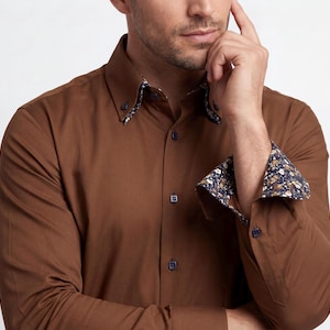 May include: A long-sleeved button-down shirt in a solid brown colour. The shirt features a button-down collar and cuffs that are lined with a dark blue fabric displaying a white and yellow floral pattern. The buttons are dark blue and square-shaped.