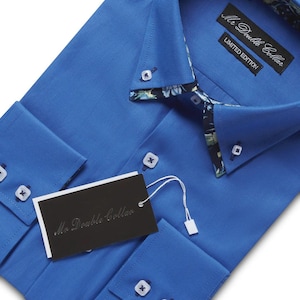 Puede incluir: Camisa azul brillante con un estampado floral en el cuello y los puños. La camisa tiene una etiqueta que dice "Mr. Double Collar" y "Limited Edition". Tiene botones blancos y está cuidadosamente doblada.