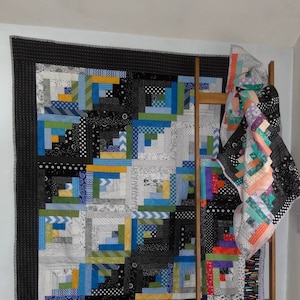 Puede incluir: Una colcha negra y blanca con detalles azules, verdes y amarillos está colgada en una pared blanca. Una escalera de madera se apoya en la pared con una colcha colorida sobre ella.