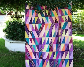 Könnte beinhalten: Ein farbenfroher Quilt mit einem geometrischen Muster aus Dreiecken in den Farben Lila, Rosa, Gelb, Orange und Blau. Der Quilt hängt an einer Wäscheleine in einem Garten.