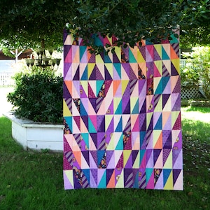 Könnte beinhalten: Ein farbenfroher Quilt mit einem geometrischen Muster aus Dreiecken in den Farben Lila, Rosa, Gelb, Orange und Blau. Der Quilt hängt an einer Wäscheleine in einem Garten.