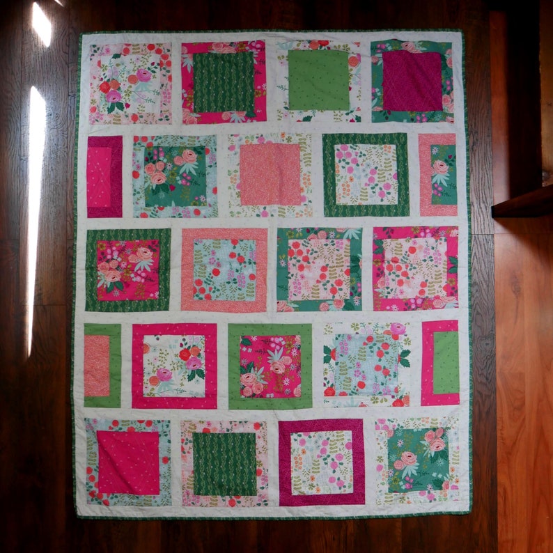 K&ouml;nnte beinhalten: Ein handgefertigter Quilt mit Patchwork-Design. Der Quilt zeigt Quadrate mit verschiedenen Blumenmustern in Rosa, Gr&uuml;n und Wei&szlig;, eingerahmt von einem wei&szlig;en Rand. Der Quilt ist etwa 152 cm lang und 102 cm breit.