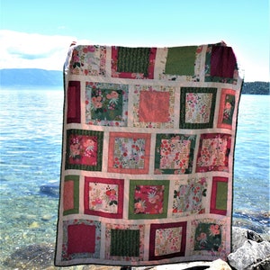 K&ouml;nnte beinhalten: Ein farbenfroher Patchwork-Quilt mit floralem Muster und quadratischen Bl&ouml;cken in Rosa, Gr&uuml;n und Wei&szlig;. Der Quilt wird im Freien mit blauem Himmel und Wasser im Hintergrund pr&auml;sentiert. Der Quilt ist ca. 152 cm x 178 cm gro&szlig;.