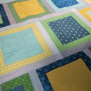 K&ouml;nnte beinhalten: Ein Patchwork-Quilt mit geometrischem Design. Der Quilt zeigt Quadrate in verschiedenen Farben, darunter Gelb, Gr&uuml;n, Blau und Grau. Einige Quadrate haben gemusterte Stoffe, w&auml;hrend andere einfarbig sind. Der Quilt ist mit wei&szlig;em Faden zusammengen&auml;ht.