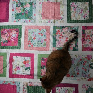 K&ouml;nnte beinhalten: Ein Patchwork-Quilt mit einem Blumenmuster in Rosa-, Gr&uuml;n- und Blaut&ouml;nen. Der Quilt hat quadratische Bl&ouml;cke mit verschiedenen Blumendesigns und einfarbigen R&auml;ndern. Eine braune und schwarze Katze liegt auf dem Quilt.