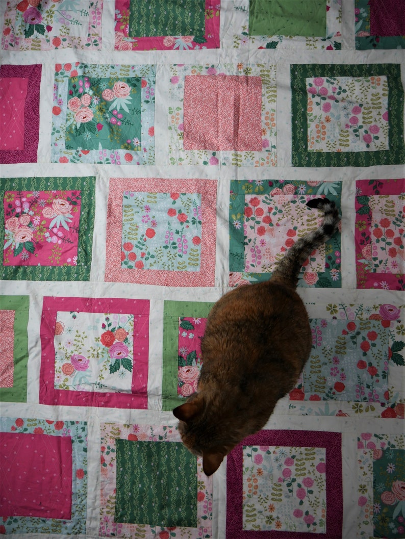 K&ouml;nnte beinhalten: Ein Patchwork-Quilt mit einem Blumenmuster in Rosa-, Gr&uuml;n- und Blaut&ouml;nen. Der Quilt hat quadratische Bl&ouml;cke mit verschiedenen Blumendesigns und einfarbigen R&auml;ndern. Eine braune und schwarze Katze liegt auf dem Quilt.