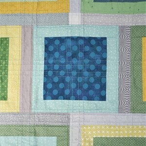 K&ouml;nnte beinhalten: Nahaufnahme eines Patchwork-Quilts mit geometrischem Design. Der Quilt zeigt Quadrate und Rechtecke in Blau-, Gr&uuml;n-, Gelb- und Graut&ouml;nen. Das mittlere Quadrat ist dunkelblau mit helleren blauen Punkten. Der Quilt ist mit wei&szlig;em Faden gen&auml;ht.
