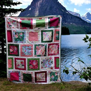 K&ouml;nnte beinhalten: Ein farbenfroher Patchwork-Quilt mit floralem Muster, drau&szlig;en mit Berg- und Seehintergrund pr&auml;sentiert. Der Quilt zeigt Quadrate aus rosa, gr&uuml;nen und wei&szlig;en Stoffen mit floralen Designs. Der Quilt wird hochgehalten.