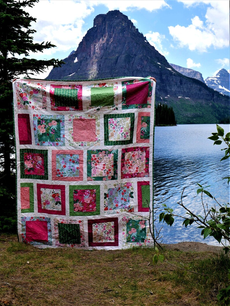 K&ouml;nnte beinhalten: Ein farbenfroher Patchwork-Quilt mit floralem Muster, drau&szlig;en mit Berg- und Seehintergrund pr&auml;sentiert. Der Quilt zeigt Quadrate aus rosa, gr&uuml;nen und wei&szlig;en Stoffen mit floralen Designs. Der Quilt wird hochgehalten.