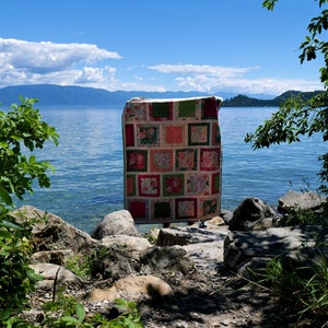 K&ouml;nnte beinhalten: Eine handgefertigte Steppdecke mit einem Patchwork-Design mit Quadraten aus floralen und unifarbenen Stoffen in Rosa-, Gr&uuml;n- und Rott&ouml;nen. Die Steppdecke wird im Freien mit einem See und Bergen im Hintergrund ausgestellt.