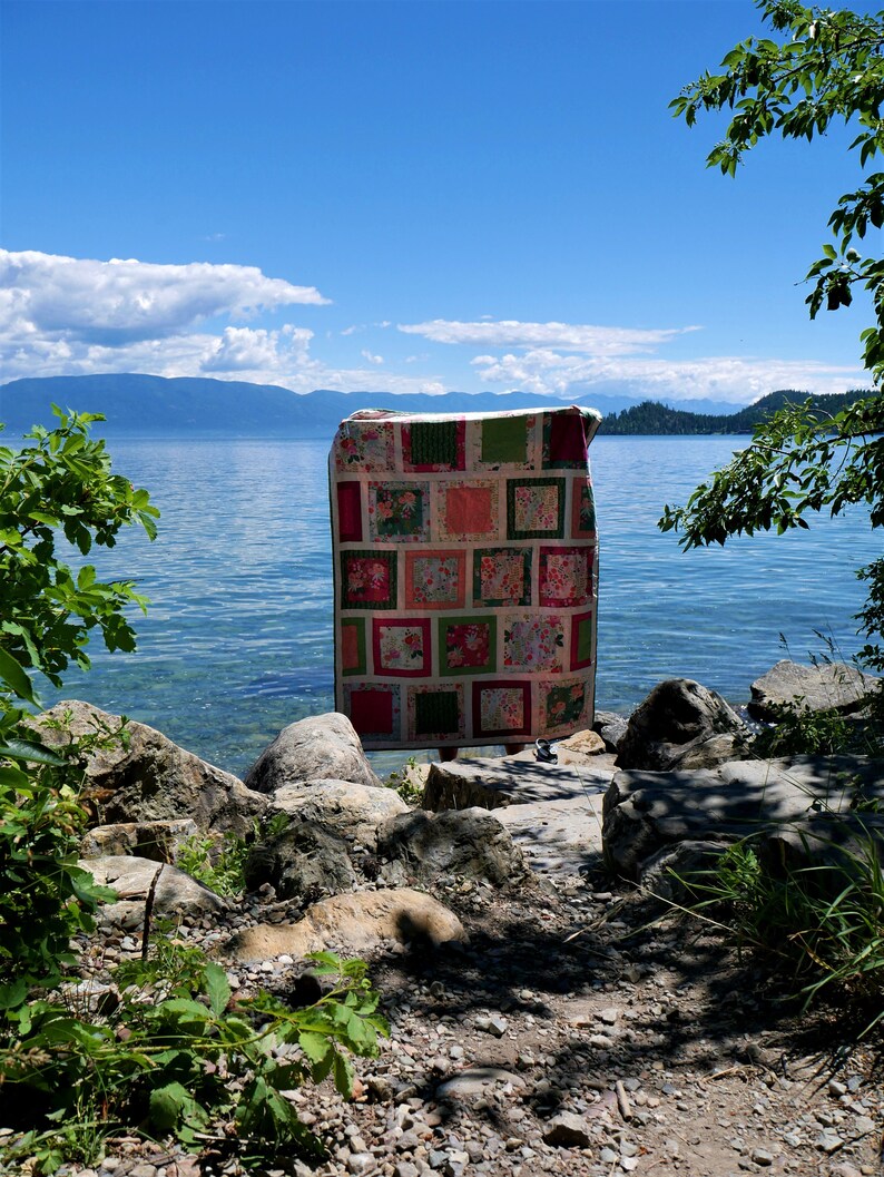 K&ouml;nnte beinhalten: Eine handgefertigte Steppdecke mit einem Patchwork-Design mit Quadraten aus floralen und unifarbenen Stoffen in Rosa-, Gr&uuml;n- und Rott&ouml;nen. Die Steppdecke wird im Freien mit einem See und Bergen im Hintergrund ausgestellt.