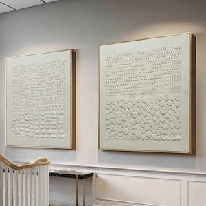 May include: Two square wall art pieces featuring off-white textured surfaces with raised, repeating dot and circle patterns. Each canvas is framed in a thin gold metal border, displaying a monochromatic design with tactile, three-dimensional elements.