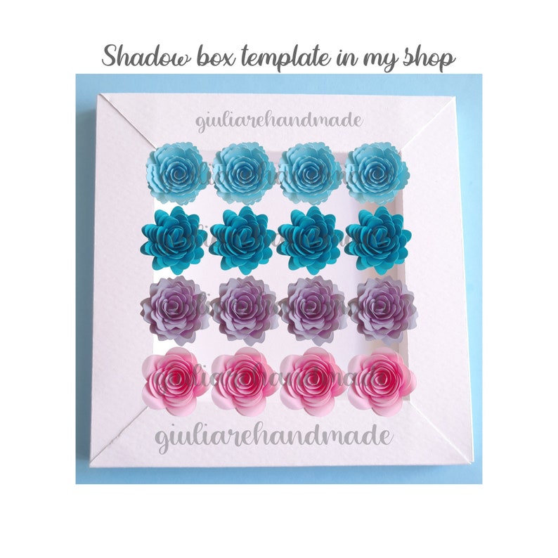 May include: A white shadow box template with rows of paper flowers in shades of blue, teal, purple, and pink. The text "Shadow box template in my shop" and "giuliarehandmade" are visible.