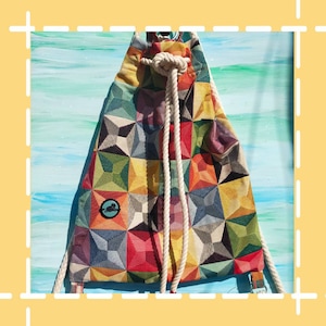 May include: A colorful drawstring backpack with a geometric pattern in shades of red, green, yellow, and blue. The bag features a white rope drawstring closure and a small circular emblem. The background is a light blue and green painted surface.