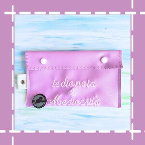 Può includere: Una pochette con zip di colore lavanda con cuciture bianche e due bottoni bianchi. La pochette è ricamata con il testo "tedio noia & mediocrità" in bianco.