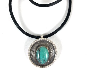 May include: A silver pendant necklace with a turquoise stone. The pendant has a circular design with a rope-like border and etched details. The necklace has a black cord.