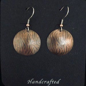May include: A pair of handcrafted copper earrings with a woodgrain pattern. The earrings are round and have a silver hook.