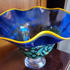 May include: A blue glass bowl with a yellow rim and a green and blue floral design. The bowl has a ruffled edge and a clear glass base.