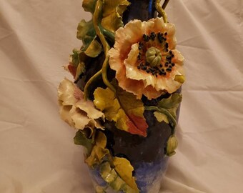 May include: A blue and white ceramic vase with a raised floral design. The vase is decorated with three dimensional flowers and leaves in shades of yellow, green, pink, and orange.