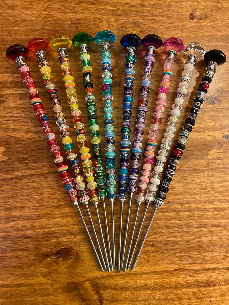 May include: A set of 12 colorful beaded wands with crystal tops. Each wand features a different color scheme and pattern of beads. The wands are made of metal and have a silver finish.