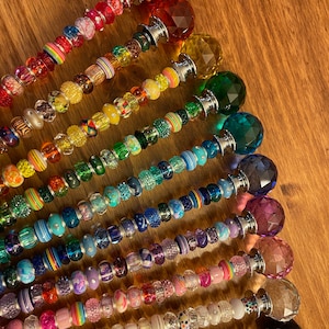 May include: A set of 14 colorful beaded wands with crystal tops. Each wand features a different color scheme and a variety of beads, including glass, metal, and plastic. The wands are arranged in a rainbow pattern, with the red wand at the top and the black wand at the bottom.