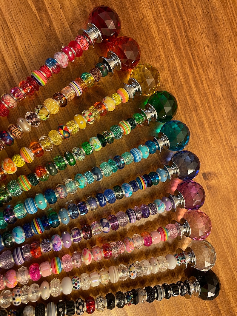May include: A set of 14 colorful beaded wands with crystal tops. Each wand features a different color scheme and a variety of beads, including glass, metal, and plastic. The wands are arranged in a rainbow pattern, with the red wand at the top and the black wand at the bottom.